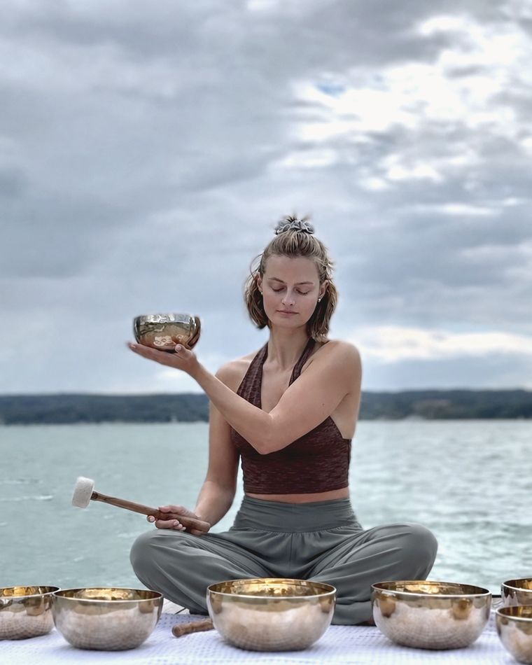 Toya sitting cross-legged on a wooden jetty by a lake, eyes closed, holding a singing bowl, with more bowls in front of her
